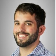 Lucas Roach, Xcel Energy. A man with short dark hair and a beard smiles at the camera. He is wearing a blue and white checkered shirt and is posed in front of a plain, light gray background.