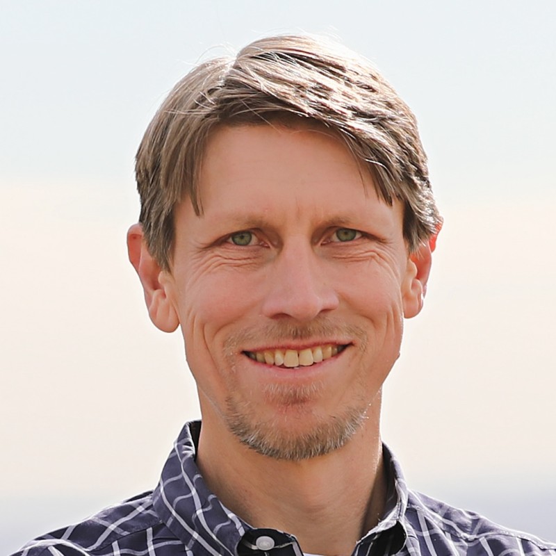 McGee Young, WattCarbon. A man with short light brown hair and a trimmed goatee, smiling, wearing a blue and white checkered shirt, standing outdoors with a pale sky in the background.