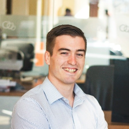 A man in a checkered shirt smiles while standing in a modern office with glass walls in the background.