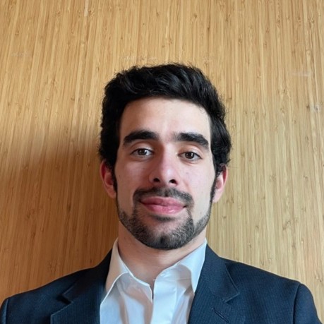 Nick Scaramuzzino, Orange and Rockland. A man with dark hair and a trimmed beard, wearing a white shirt and dark blazer, poses in front of a wooden textured background.