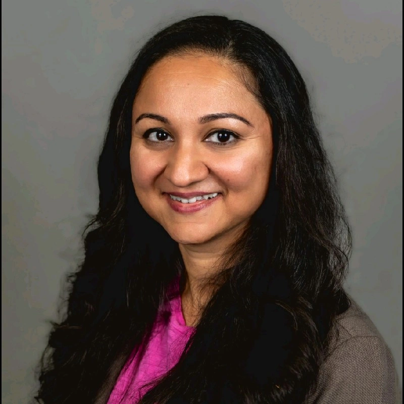 Nikhila Datta, BGE. A woman with long dark hair and brown eyes smiles at the camera. She is wearing a pink top and a brown jacket, posed in front of a plain gray background.