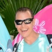 Robert Johnson, Tampa Electric Company. A smiling person wearing sunglasses and a light blue shirt stands in front of palm leaves, with colorful surfboards—one pink with a white hibiscus design—visible behind them.