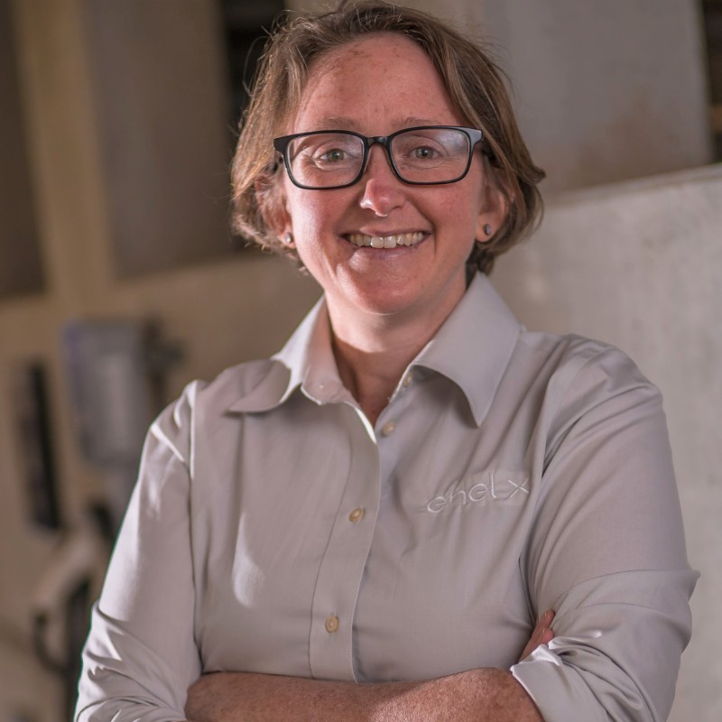 Sally Griffith, Resource Innovations. A person with short brown hair and glasses smiles while standing with arms crossed, wearing a light gray button-up shirt with chelX embroidered on it. The background is softly blurred indoors.