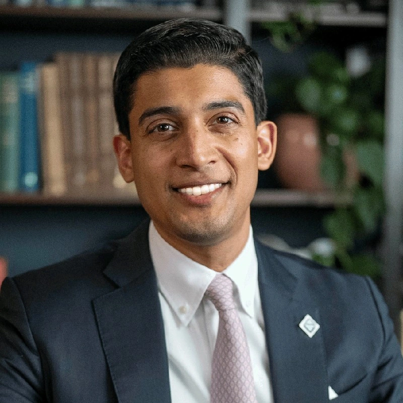 Varun Sivaram, Emerald AI. A man in a suit and tie smiles while sitting indoors, with books and plants on shelves in the background.