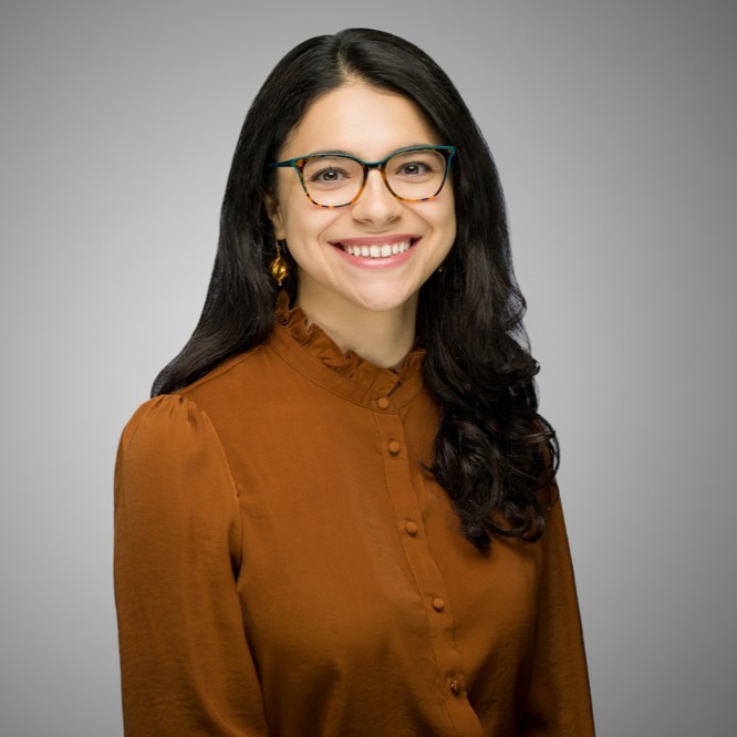 A woman with long, dark hair wearing glasses and a brown blouse smiles at the camera. She is posed in front of a plain gray background.