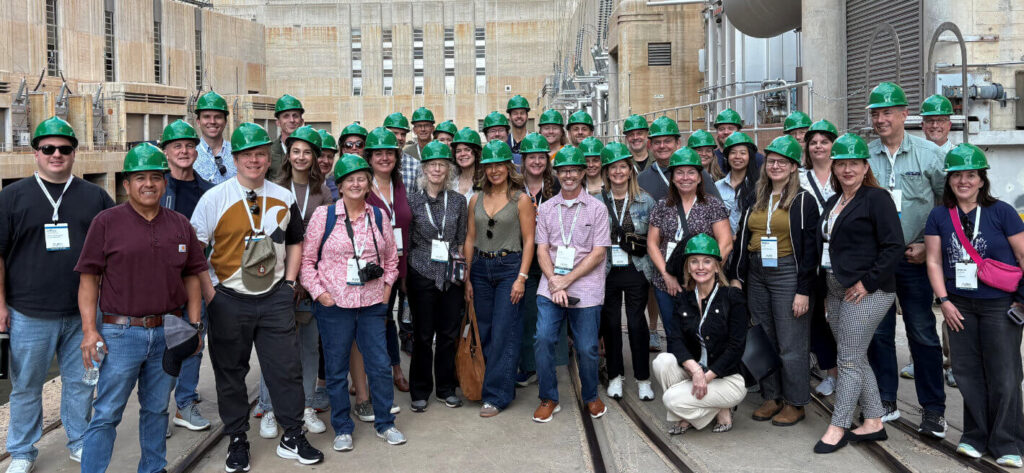 A large group of people wearing green hard hats pose together for a photo at an industrial or construction site.