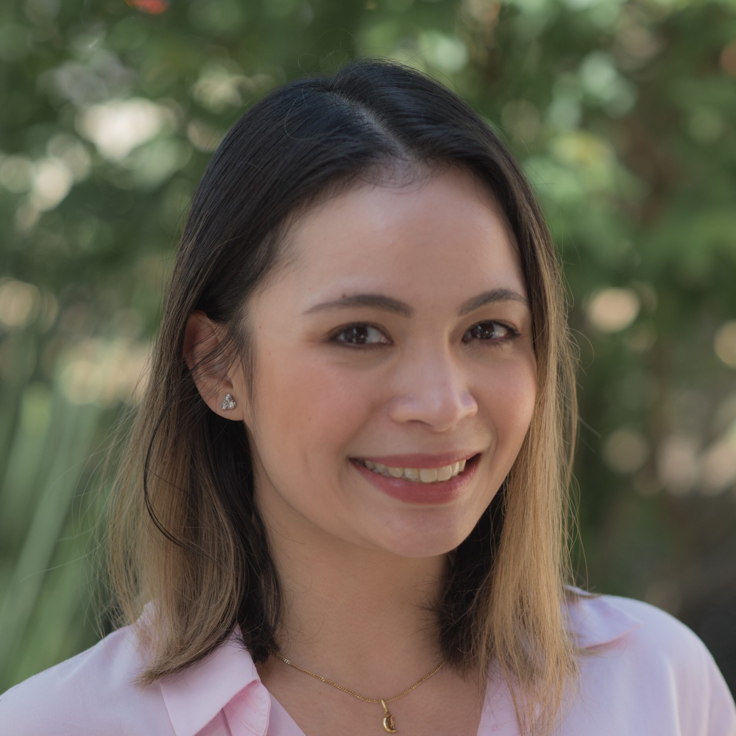 Woman with shoulder-length hair, wearing a light pink blouse, smiling outdoors with greenery in the background.