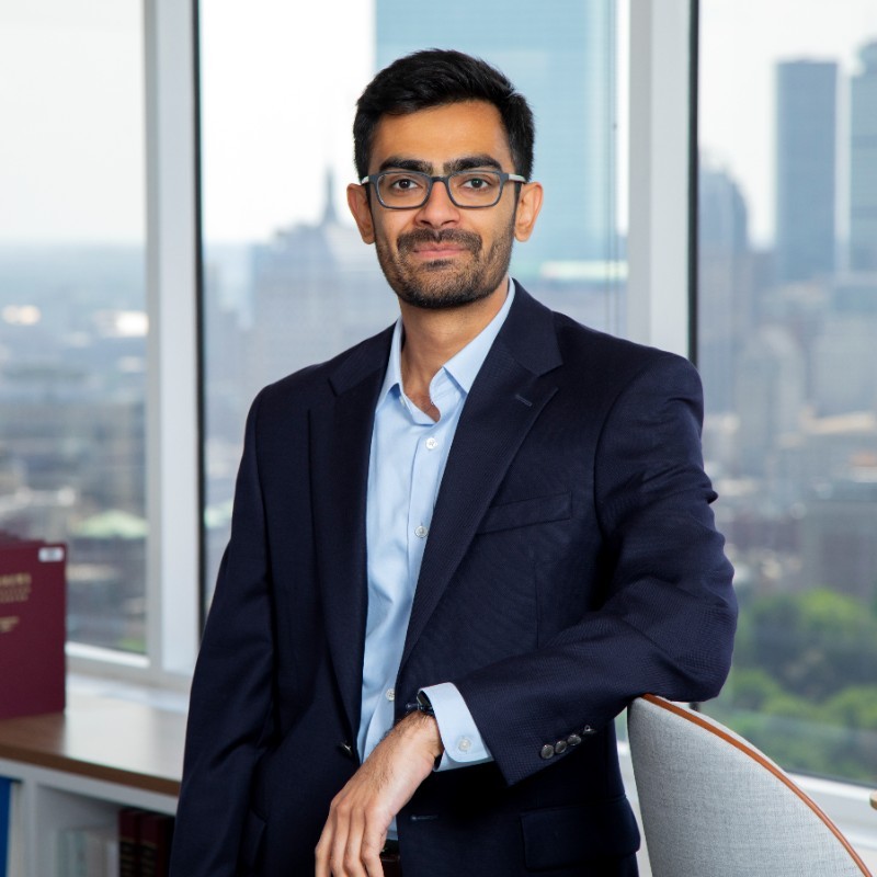 Akhilesh Ramakrishnan, The Brattle Group. A man in a navy suit and glasses sits in an office with large windows overlooking a cityscape.
