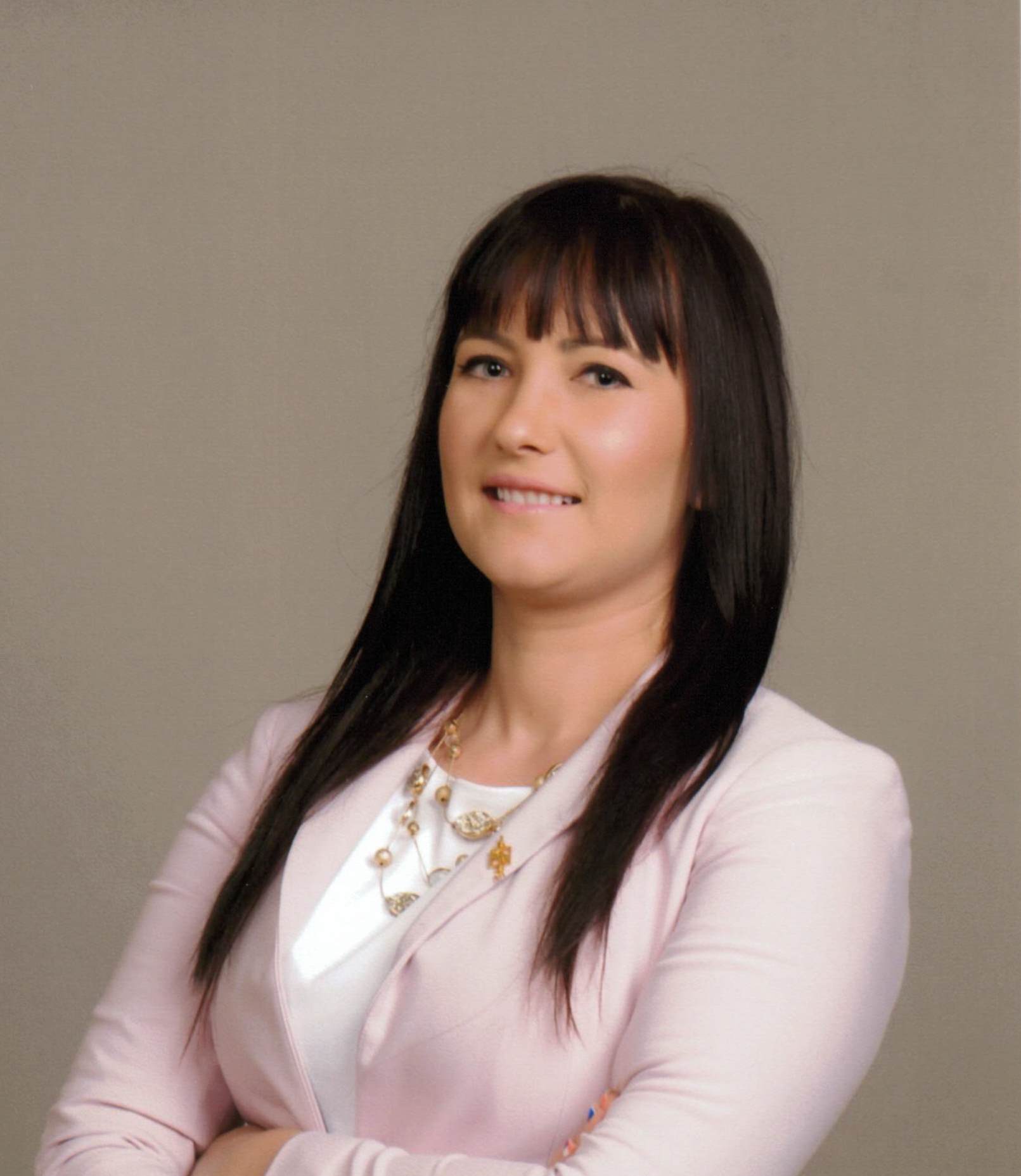 Woman with straight dark hair, wearing a light pink blazer and white top, smiling with arms crossed against a plain light background.