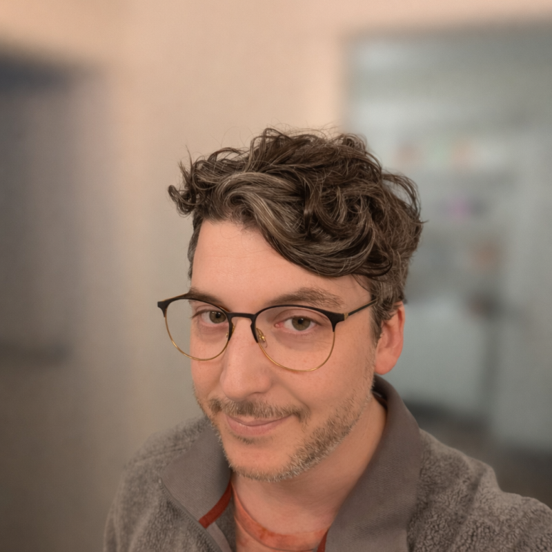 Anthony Ionno, IESO. Man with wavy brown hair, glasses, and a gray zip-up sweater smiles in a softly blurred indoor setting.