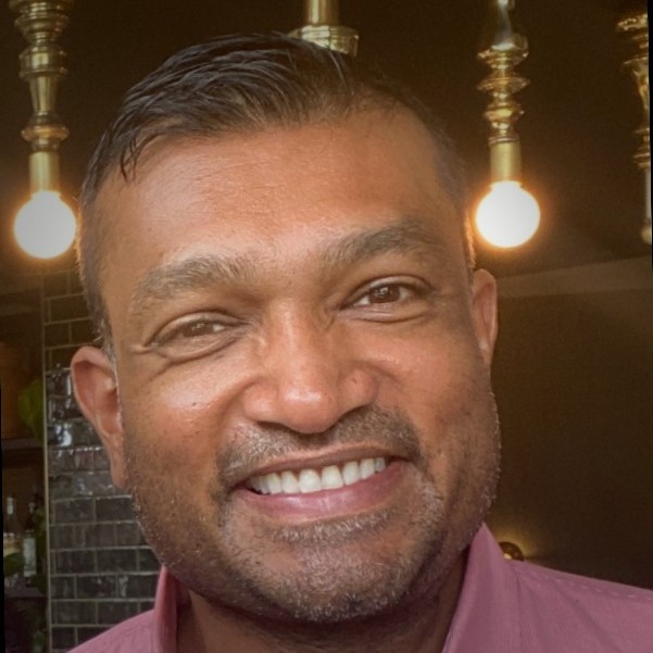 Divesh Gupta, ev.energy. Man smiling indoors with light bulbs and dark tiles in the background, wearing a pink collared shirt.