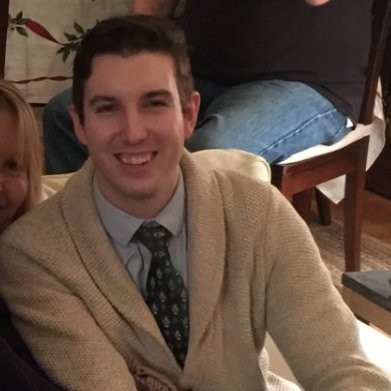 A man in a beige cardigan and patterned tie smiles while sitting indoors on a couch with another person partially visible.