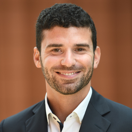 Man in a suit smiling in front of a blurred, brownish background. He has short dark hair and a trimmed beard.