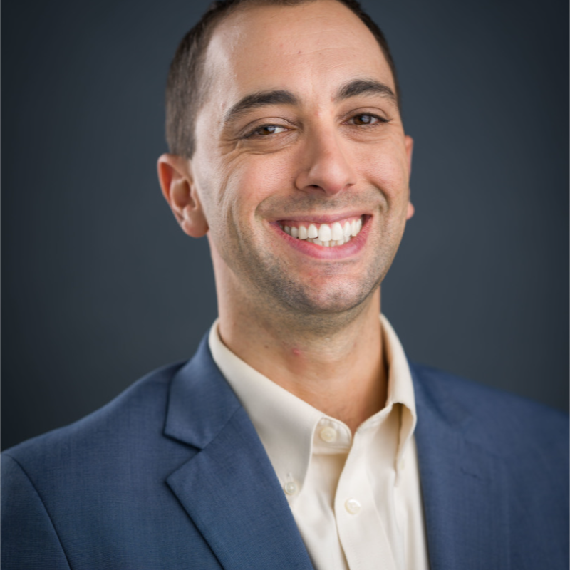 Jed Cohen, Salt River Project. Smiling man in a blue suit jacket and cream shirt, posing in front of a dark, plain background.