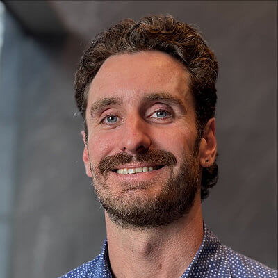 Ken Norris, Dominion Energy. Smiling man with curly brown hair and mustache, wearing a blue patterned shirt, standing in front of a gray background.