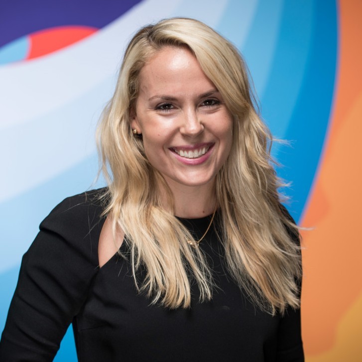 Liz Faralli, Treehouse. A woman with long blonde hair wearing a black top smiles in front of a colorful abstract background.