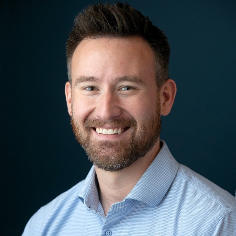 Luke Beckett, E Source. Man with short brown hair and beard, wearing a light blue shirt, smiling against a dark blue background.