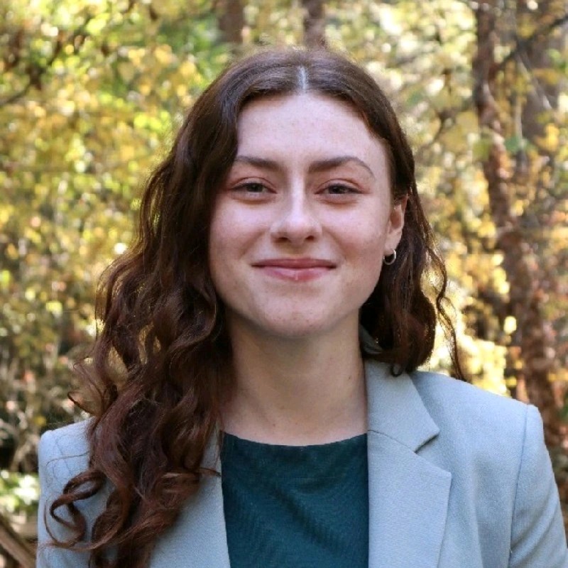 Young woman with long curly hair and a light blazer smiles outdoors, with green and yellow trees in the background.