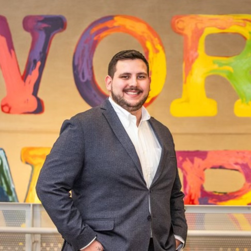A man in a suit stands smiling in front of a colorful wall with large painted letters.