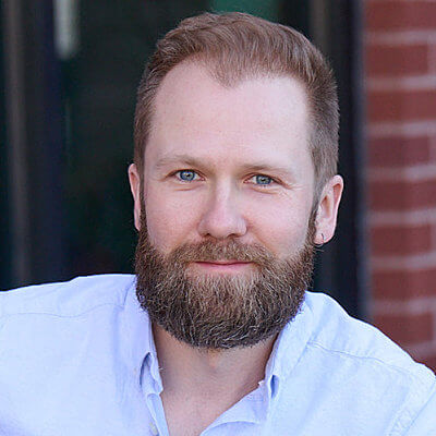 Bearded man with light brown hair and blue eyes, wearing a light blue shirt, smiling in front of a brick wall.