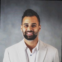 Smiling man with dark hair and beard wearing a light-colored blazer and white shirt, posed against a gray background.