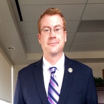 Nicholas Bennett, CPS Energy. A man in a suit and striped tie stands indoors, smiling slightly, with office ceiling tiles visible above.
