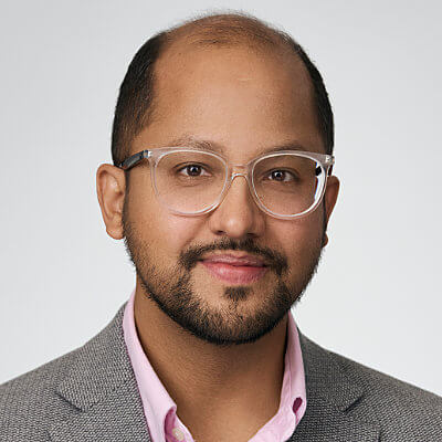 A man with glasses, a beard, and a mustache, wearing a gray blazer and pink shirt, smiles at the camera against a light background.