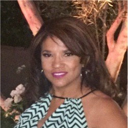 Robyne Black, Ameren. Woman smiling outdoors at night, wearing a sleeveless zigzag-patterned dress with dark brown, wavy hair and pink lipstick.