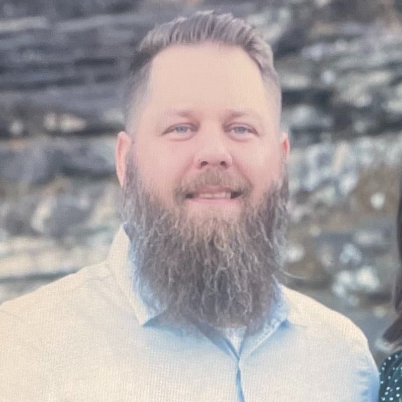 Shane King, OG&E. Man with a full beard and light-colored shirt smiles in front of a stone wall background.