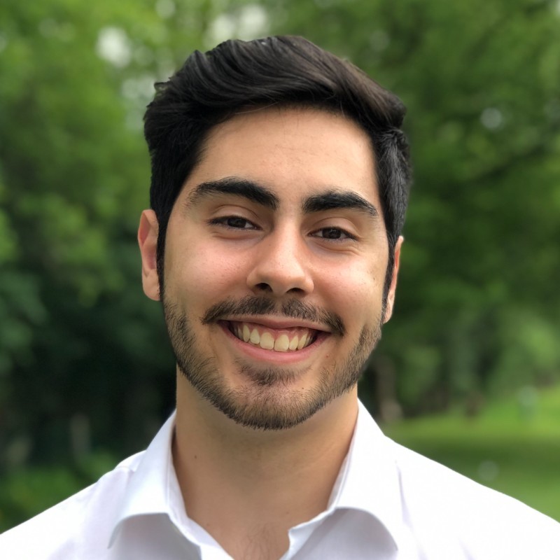 Simitrio Arellano, ComEd. Young man with dark hair and beard smiling, wearing a white shirt, standing outdoors with green trees in the background.