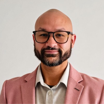 Tyson Brown. Bald man with glasses and beard, wearing a pink blazer and light shirt, smiling in front of a plain background.