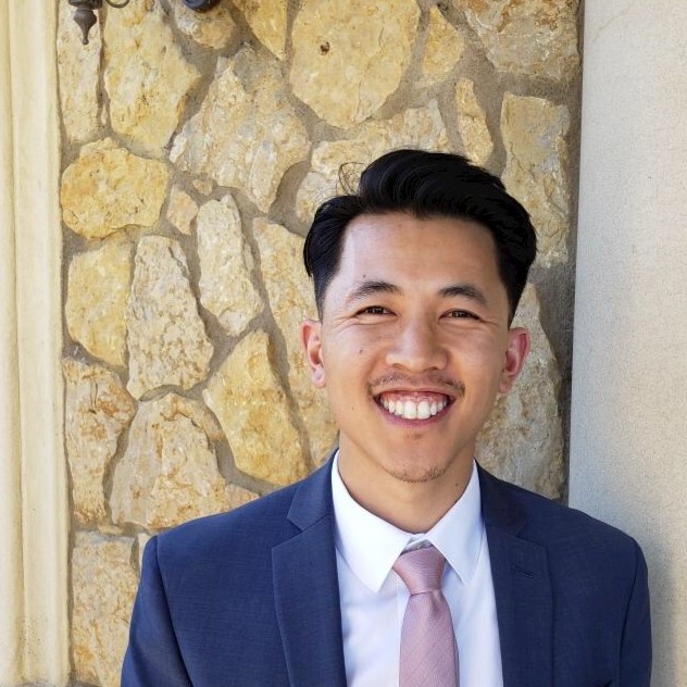 Yao Chao, Puget Sound Energy. Man in a blue suit and light pink tie smiles while standing in front of a stone wall.