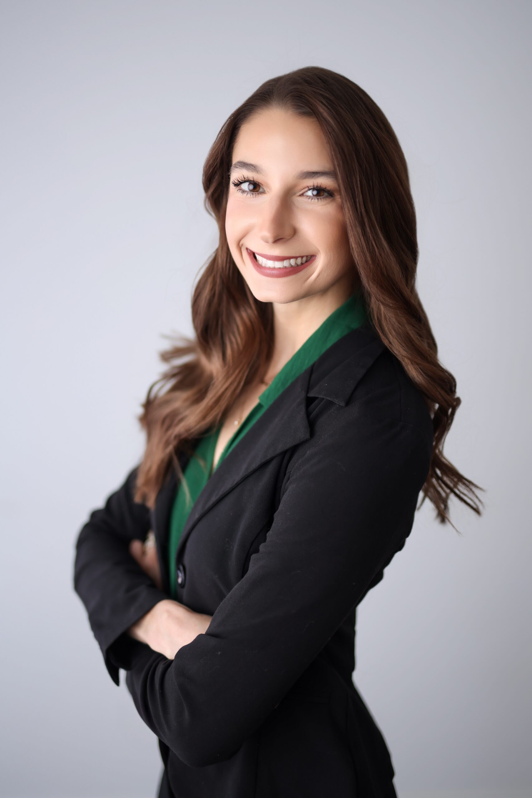 Woman with long brown hair in a black blazer and green shirt smiles with arms crossed against a plain light background.