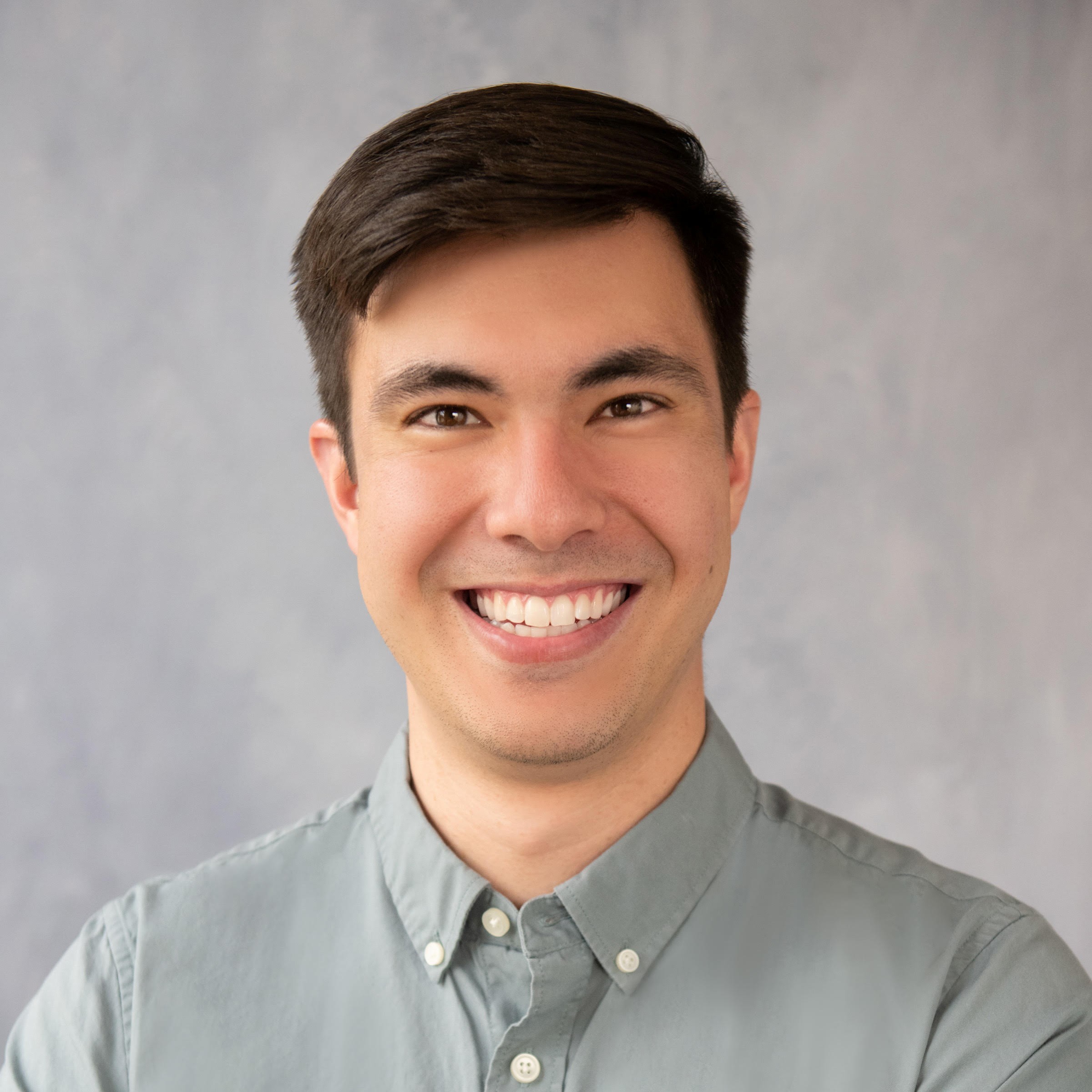 Smiling man with short dark hair wearing a light gray collared shirt, posing in front of a soft gray background.