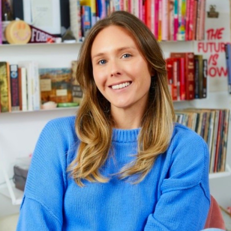 Brittney Winters-Gullo, EnergyHub. Woman with long hair in a blue sweater smiles, sitting in front of a colorful bookshelf filled with books and decor.