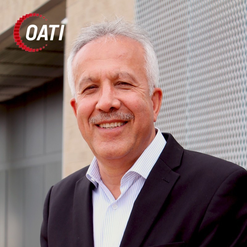 Smiling man in a suit stands outdoors near a metal wall with an OATI logo in the upper left corner.