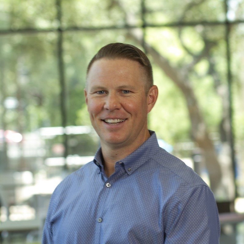 Smiling man in a blue button-up shirt stands indoors with large windows and trees in the background.