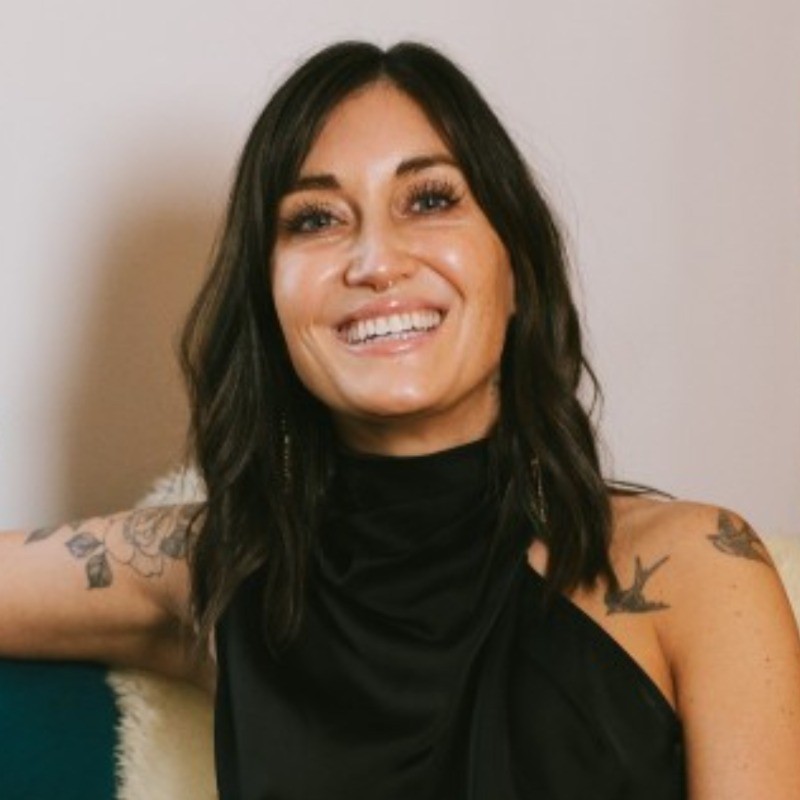 A woman with dark hair, wearing a sleeveless black top, smiles while sitting on a couch. Tattoos are visible on her arms.