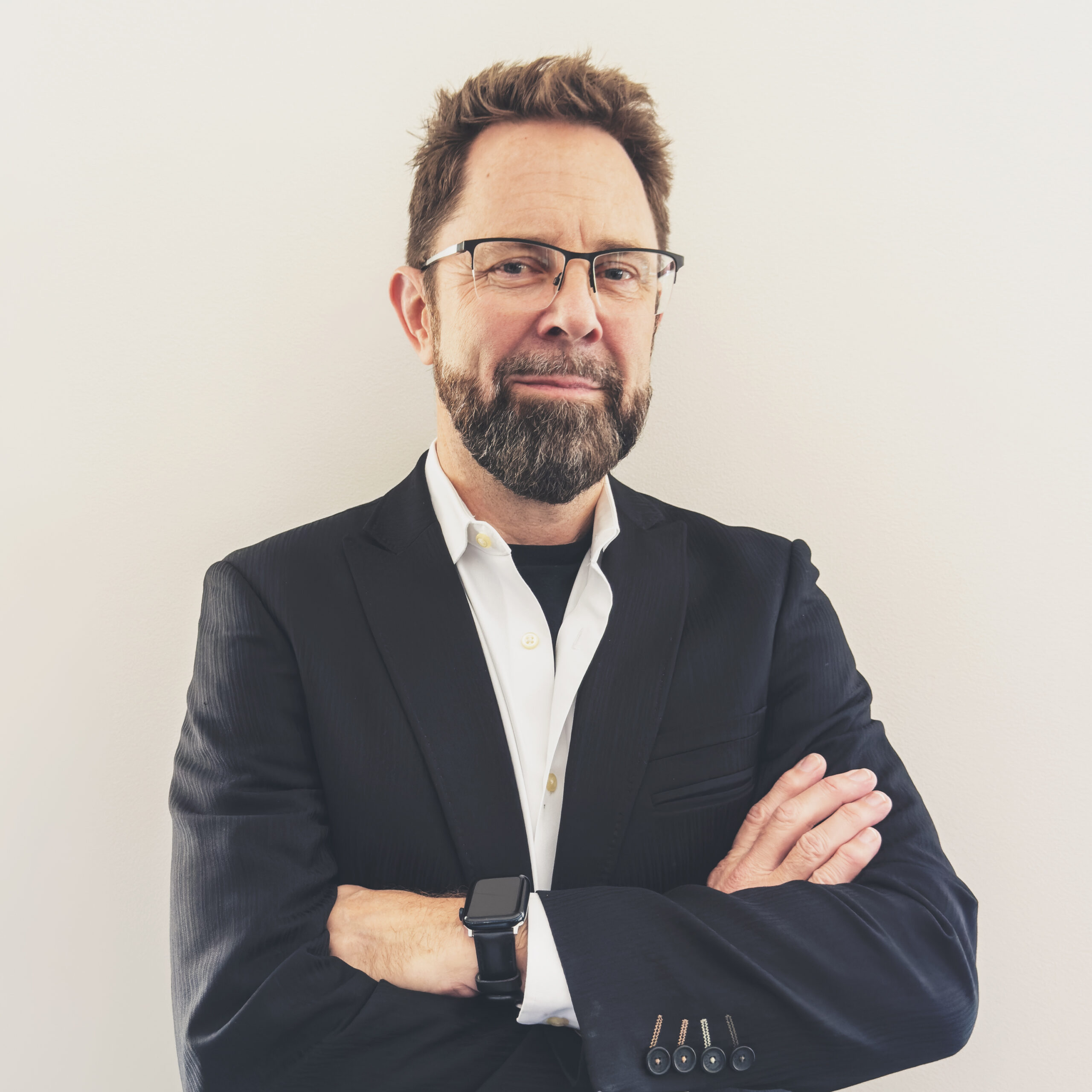 Man with glasses and beard wearing a suit and smartwatch, standing with arms crossed against a light background.