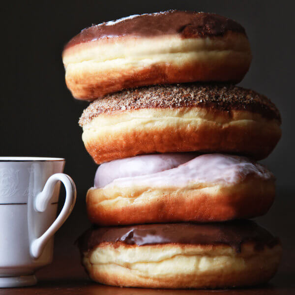 A stack of four assorted frosted donuts next to a white teacup on a dark background.
