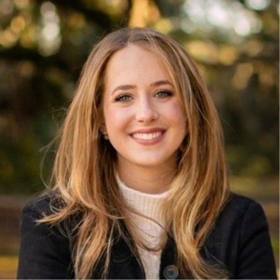 Alexandra Heuer, Entergy. Smiling woman with long blonde hair, wearing a black jacket and white sweater, standing outdoors with trees in the background.