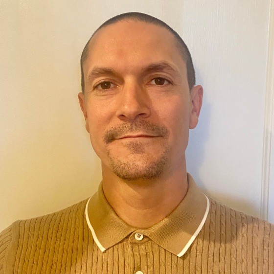 Felipe Godinez, Olivine. Man with short dark hair and a trimmed goatee wearing a tan collared shirt stands in front of a white wall, looking at the camera.