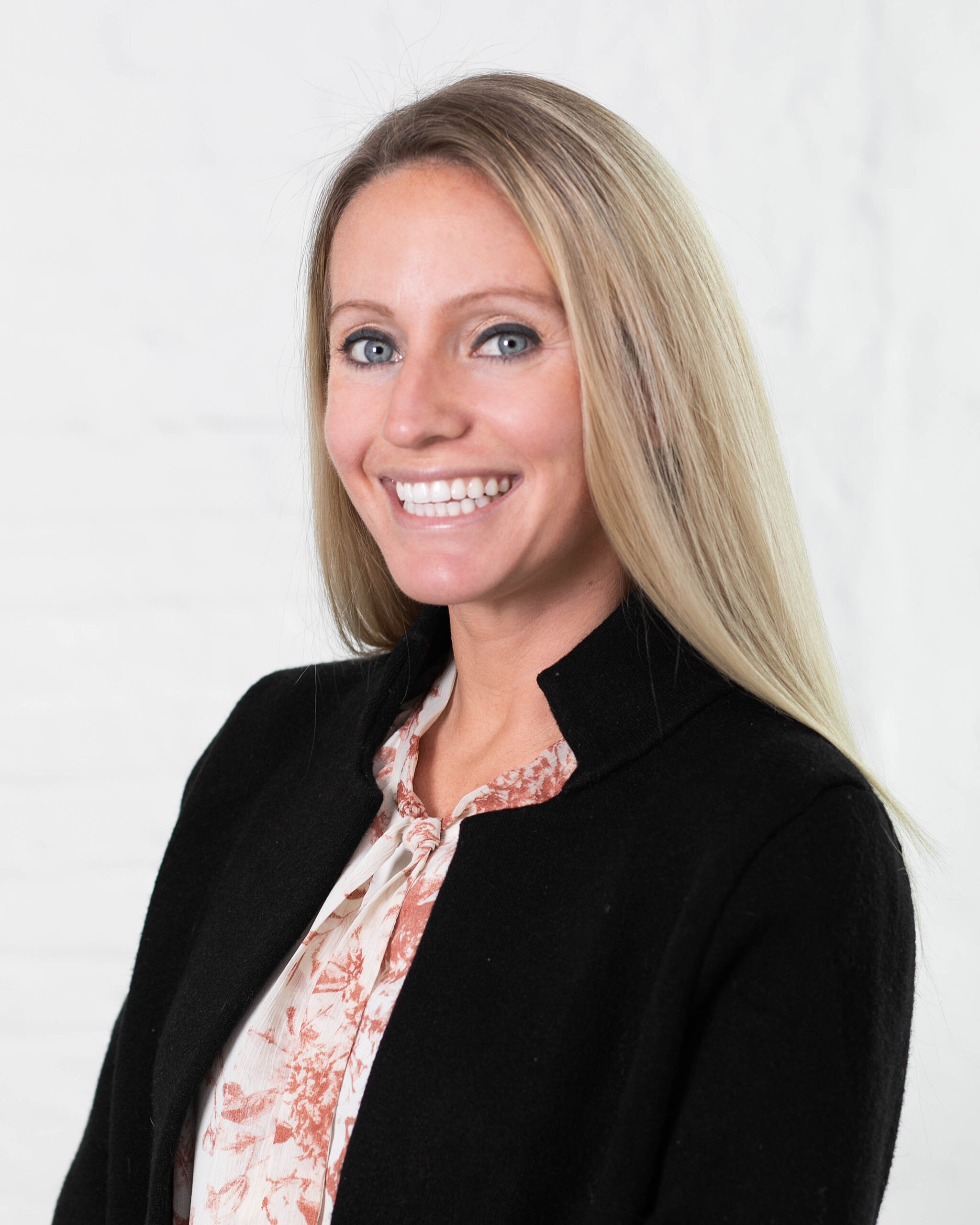 Smiling woman with long blonde hair, wearing a black blazer and patterned blouse, posed against a light background.