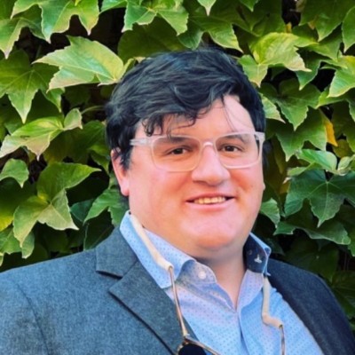 Maxwell Burke-Scoll, EnergyHub. A man wearing glasses and a blazer posing in front of a leafy green wall.